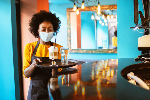 istockphoto-1300423378-612×612 Beautiful Afro-American woman works in a cafe as a waitress with a protective mask on her face and holds a coffee latte on a tray.
